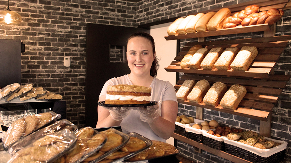 Owner at the store counter holding a cake and surrounded by baked goodies